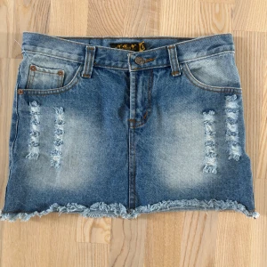 Blå jeanskjol med slitna detaljer - Snygg blå jeanskjol från M&N Jeans med slitna detaljer framtill och baktill. Kjolen har fransig nederkant, klassiska fickor och bälteshällor. Använd endast ett fåtal gånger.