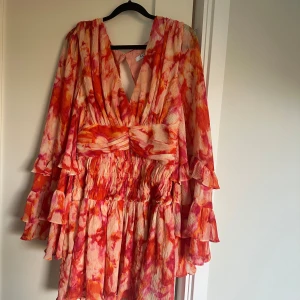 Färgstark kortklänning från NA-KD - Säljer en superfin kortklänning från NA-KD med v-ringning och volangärmar. Klänningen har ett färgstarkt blommigt mönster i rosa, orange och rött, samt smockad midja och omlott-detalj framtill. Perfekt för dig som gillar statement-plagg och snygga detaljer. Endast använd ett par gånger.