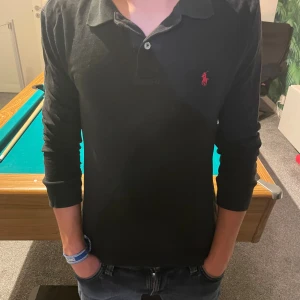 Svart långärmad pikétröja från Ralph Lauren - Svart långärmad pikétröja från Ralph Lauren med klassisk krage och knäppning framtill. Tröjan har det ikoniska röda Polo-logot broderat på bröstet och är tillverkad i mjuk bomull. Perfekt för en clean och stilren look.