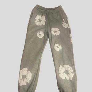 Gråa Denim Tears mjukisbyxor med vita blommor - Gråa Denimtears mjukisbyxor med elastisk midja och snörning, dekorerade med stora vita blommönster över hela byxorna. Avslappnad passform med resår vid benslut och fickor på sidorna samt bak. Perfekta för en chill och trendig look.