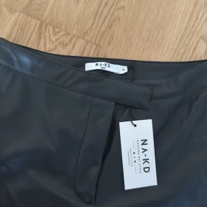 Svarta skinnbyxor från NA-KD med slits - Snygga svarta byxor i skinnimitation från NA-KD. De har en tight passform, hög midja och slitsar nertill på benen för en trendig look. Perfekta att styla med sneakers eller boots. Materialet är syntetiskt och ger en cool vibe.