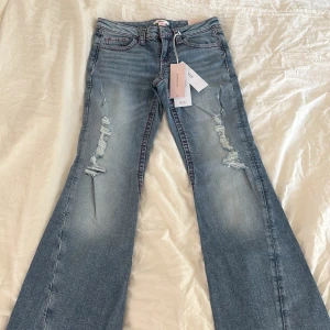 Bootcut jeans från Gina Tricot, stl 158 - Bootcut jeans i ljusblå denim från Gina Tricot med slitningar framtill och broderade bakfickor i rosa och rött. Snygga kontrastsömmar och klassisk femficksmodell. Perfekta för dig som gillar en cool och avslappnad stil.