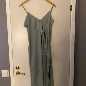 Ljusgrå långklänning med volang - Supersnygg ljusgrå långklänning med smala axelband och v-ringad hals. Klänningen har en asymmetrisk volangdetalj framtill som ger en cool och feminin vibe. Materialet är mjukt och faller snyggt längs kroppen, perfekt för dig som gillar stilrena och enkla plagg med en twist.❤️Frakt ingår inte 