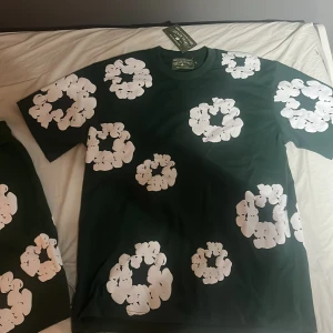 Grön blommig t-shirt och shorts från Denim Tears - Snygg grön t-shirt från Denim Tears med stora vita blommiga tryck över hela tröjan. Klassisk passform med rund hals och korta ärmar. Perfekt statement-plagg för dig som vill sticka ut med cool design och streetwear-vibe. Kommer också med shortsen om man vill ha. Size L på shortsen och M på t shirten men passar L. Så L på Båda. Priset kan diskuteras. Har inte använts något helt nya.