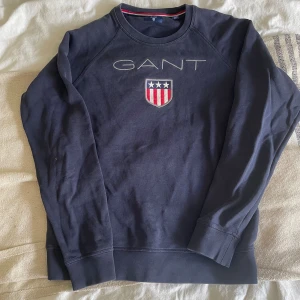 Mörkblå sweatshirt från GANT - Mörkblå sweatshirt från GANT med broderad logga och sköld på bröstet. Tröjan har rund hals, långa ärmar och ribbade muddar. Klassisk och stilren design med vita och röda detaljer i loggan. Perfekt för dig som gillar preppy stil.