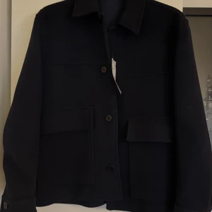 Svart overshirt med stora fickor S - Stilren svart overshirt med klassisk krage och stora fickor framtill. Jackan har knäppning med svarta knappar och raka ärmar. Perfekt för lager-på-lager och enkel att matcha med olika outfits.