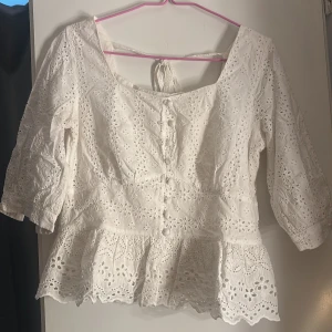 Vit broderad blus med volang - Superfin vit blus med broderade blommor och volang nertill. Blusen har trekvartsärm, knappar framtill och knytning i ryggen. Köpt i Thailand den var då för litet för mig vid brösten, det står att det är storlek L men det stämmer inte är typ S eller xs