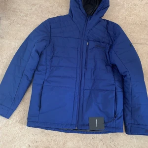 Blå pufferjacka Peak Performance - Blå pufferjacka från Peak Performance med huva och dragkedja framtill. Jackan har quiltad design, en bröstficka med dragkedja och diskret logga på bröstet. Perfekt för kalla dagar och riktigt snygg färg som sticker ut.