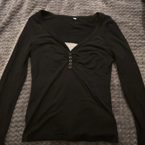 Svart långärmad topp med knappar - Svart långärmad topp med djup v-ringning och sex dekorativa knappar framtill. Toppen har en snygg rynkning vid bysten och en vit spetsdetalj i urringningen som ger en extra touch. Mjuk och stretchig, perfekt till jeans eller kjol.