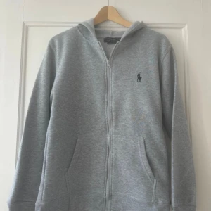 Grå hoodie från Polo Ralph Lauren - Snygg ljusgrå hoodie från Polo Ralph Lauren med dragkedja och klassisk logga på bröstet. Tröjan har huva, kängurufickor och är i en slim fit-modell. Perfekt för chill dagar eller när du vill ha en clean och enkel look.