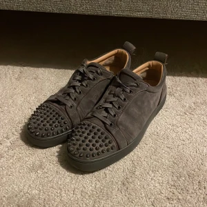 Christian Louboutin gråa med nitar - Säljer ett par gråa sneakers från Christian Louboutin i mocka med ikoniska röda sulor. Skorna har rund tå, snörning och coola gråa nitar på tån för en edgy look. Insidan är beige och sulan är platt. Perfekta för dig som vill sticka ut, storlek 40-41