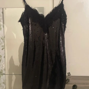 Svart paljettklänning med spets - Supersnygg svart kort klänning med paljetter och spetsdetaljer vid halsringning och nederkant. Justerbara tunna axelband för perfekt passform. Perfekt för dig som vill sticka ut med en glittrig look och älskar detaljer som spets och paljetter. Klänningen är helt ny.