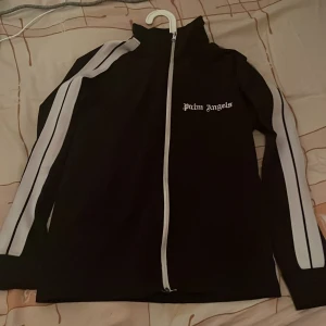 Svart track jacket från Palm Angels - Svart track jacket från Palm Angels med vita ränder längs ärmarna och vit dragkedja framtill. Jackan har hög krage och logga broderad på bröstet. Perfekt för en sportig och trendig streetwear-look har även fickor som är dragkedjor.