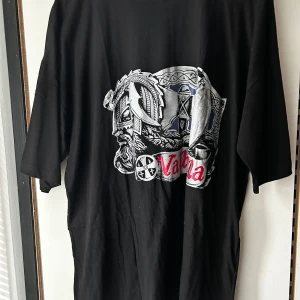 Svart Valhalla t-shirt med vikingatryck - Cool svart t-shirt med stort tryck av en viking och texten 'Valhalla' i rött framtill. T-shirten har klassisk passform och korta ärmar. Materialet känns mjukt och skönt, perfekt för dig som gillar nordisk mytologi och snygga statement-plagg.