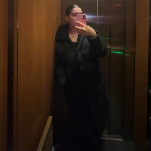 Svart lång vinterjacka  - Säljer en svart lång vinterjacka med hög krage och dragkedja framtill (finns knappar för att stänga också) . Jackan är vadderad och har en clean look som passar perfekt när det är kallt ute. Snygg och enkel att styla till olika outfits.