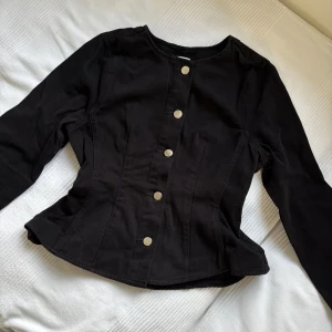Svart peplum jeansjacka strl 34 - Snygg svart peplum jeansjacka från ginatricot med markerad midja och silverfärgade knappar framtill. Jackan har lång ärm och är tillverkad i denim med en figurnära passform. Perfekt för dig som gillar en stilren och trendig look. Helt ny med prislappen, säljer pga förliten