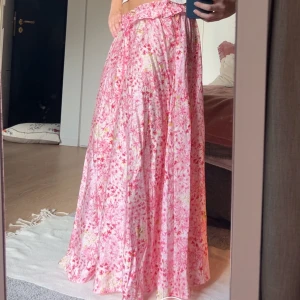 Rosa långkjol från H&M - Superfin helt ny långkjol från H&M i rosa med blommigt mönster och volangkant upptill. Kjolen har knytband i midjan och är gjord i ett lätt och luftigt material, perfekt för varma dagar. Aldrig använd. 
