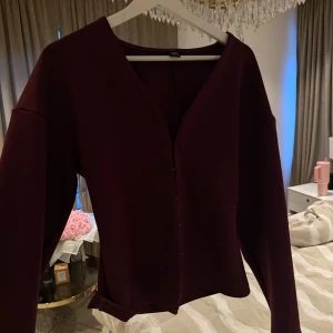 Vinröd långärmad blus med v-ringning - Säljer en stilren vinröd blus med v-ringad hals och långa ärmar. Blusen har en enkel och elegant design utan synliga detaljer, perfekt för dig som gillar en clean look. Materialet känns mjukt och följsamt mot kroppen.