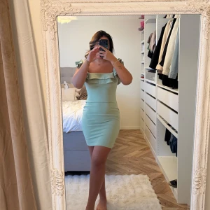 Mintgrön kort klänning med volang - Superfin mintgrön kort klänning med volangdetaljer upptill och offshoulder-stil. Klänningen är figurnära och har en snygg, tight passform som framhäver siluetten. Perfekt för dig som vill sticka ut med en fräsch färg och trendiga detaljer.