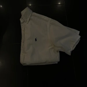 Vit kortärmad skjorta från Ralph Lauren - Vit kortärmad skjorta från Ralph Lauren med klassisk krage och knäppning framtill. Skjortan har den ikoniska broderade loggan på bröstet och är tillverkad i mjuk bomull. Perfekt för en clean och stilren look.