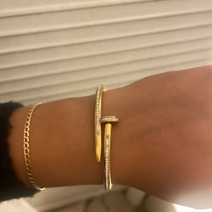 Guldarmband med stenar i spikdesign - Snyggt armband i guld- och silverfärg med en unik spikformad design. Armbandet är dekorerat med små glittrande stenar längs hela ovansidan, vilket ger en lyxig och modern känsla. Perfekt för dig som vill sticka ut med ett coolt och trendigt smycke. Tappat någon diamant annars som ny.