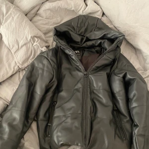 Svart vinterjacka med huva från Zara - Säljer en svart skinnjacka från Zara med huva och dragkedja framtill. Jackan har en clean look med diskreta sömmar och två fickor med dragkedja. Perfekt för dig som gillar en stilren vibe och vill ha något som sticker ut lite extra.