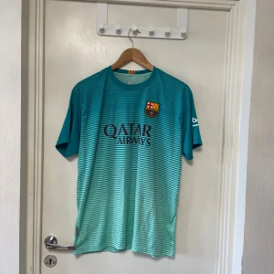 FC Barcelona turkos matchtröja L - Säljer en officiell FC Barcelona fotbollströja i turkos med tunna horisontella ränder och klubbmärke på bröstet. Tröjan har Qatar Airways-tryck på framsidan och är tillverkad i lätt, ventilerande polyester. Perfekt för dig som älskar Barca och vill sticka ut på planen.