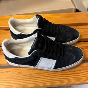 Valentino Flycrews svart/vita med nitar  - Snygga sneakers från Valentino Garavani i svart mocka och vitt skinn. Skorna har svarta snören, vit detalj på sidan och coola nitar runt hälen. 
