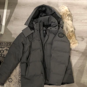  Canada Goose wyndham jacka - Säljer en grå dunjacka från Canada Goose med svart logotyp på ärmen. Modellen är Wyndham. Köpt 2018, Jackan har huva med avtagbar fuskpäls och dragkedja framtill. Perfekt för kalla dagar och har en klassisk, stilren look. Fodrad insida och flera praktiska fickor. För mer information skriv gärna. Kan även mötas upp