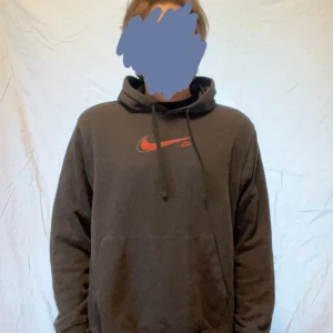 Svart Nike hoodie med röd logo - Svart hoodie från Nike med klassisk röd swoosh-logga på bröstet. Tröjan har huva med snörning och en stor magficka framtill. Tillverkad i mjuk bomullsmix, perfekt för chill dagar eller streetwear-stil. Passar M 