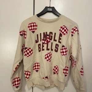 Beige jultröja med Jingle Bells-tryck - Beige sweatshirt med texten JINGLE BELLS framtill och röda julgranskulor med rutigt mönster över hela tröjan. Rund halsringning och långa ärmar. Perfekt för julens alla tillfällen!