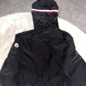 Svart vindjacka från Moncler med huva - Svart vindjacka från Moncler med huva och dragkedja framtill. Jackan har Moncler-logga på ärmen och snygga röd, vit och blå detaljer på huvkanten. Tillverkad i lätt polyester, perfekt för blåsiga dagar. Snygg och stilren design som passar till det mesta.