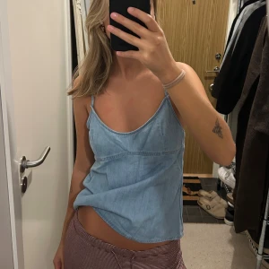 Ljusblå denim linnetopp med knyt - Säljer en ljusblå linnetopp i denim-look med tunna axelband och snygg öppen rygg med knyt. Toppen har en avslappnad passform och är perfekt för varma dagar. Enkel och stilren design som är lätt att matcha.