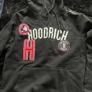 Svart hoodie från Hoodrich med patches - Svart hoodie från Hoodrich med stora broderade patches och tryck i vitt och rött framtill. Klassisk huva med snörning och känguruficka. Snygg streetwear-stil med detaljerade loggor och kontrasterande färger.