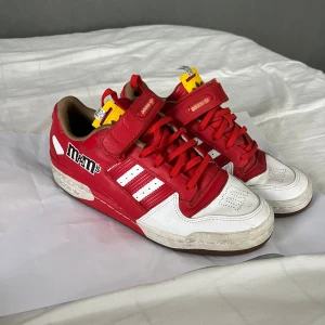 Adidas Forum Low M&M's röd/vit - Unika Adidas Forum Low sneakers i samarbete med M&M's. Skorna är röda och vita med klassiska tre ränder, snörning och kardborreband. M&M's-logga på sidan och detaljer i gult på plösen. Tillverkade i skinn och syntet för en cool och färgstark look.