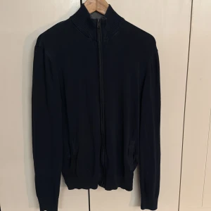 Mörkblå kofta från Pier One - Mörkblå kofta från Pier One med hel dragkedja och ribbad hög krage. Tröjan har långa ärmar, två fickor framtill och är tillverkad i mjukt material som känns skönt mot huden. Perfekt för lager-på-lager under kyliga dagar.