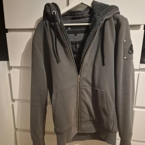 Grå hoodiejacka Moose Knuckles - Säljer en grå/grön hoodiejacka från Moose Knuckles med dragkedja, huva och mjukt foder. Jackan har två stora fickor fram, ribbade muddar och snygg logotyp på ärmen. Perfekt för dig som gillar streetwear och vill ha en stilren look. Mindre fläck på framsidan