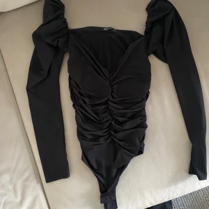 Svart långärmad body från NLY One - Snygg svart body med långa puffärmar och djup v-ringning från NLY One. Toppen har rynkade detaljer framtill som ger en cool och trendig look. Tillverkad i stretchigt material för en tight passform.