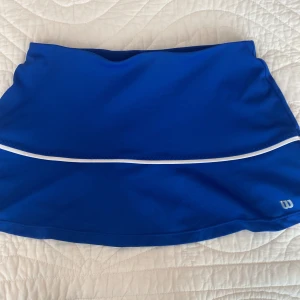 Blå tennis kjol från Wilson - Sportig blå mini skirt från Wilson med vit detalj och inbyggda shorts under. Kjolen är tillverkad i ett stretchigt syntetmaterial som passar perfekt för tennis eller träning eller om man bara vill ha en gullig mini kjol. Enkel och stilren design med diskret Wilson-logga framtill.