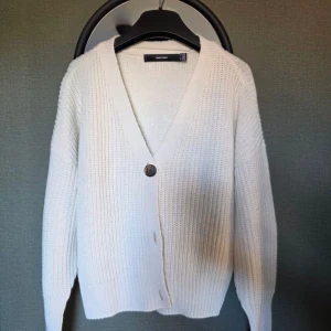 Vit stickad kofta från Vero Moda - Mysig vit stickad kofta från Vero Moda med v-ringning och en stor knapp framtill. Koftan har lång ärm och en loose passform, perfekt att slänga på sig över en t-shirt eller linne. Enkel och stilren design som funkar till det mesta.