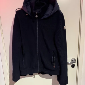 Mörkblå stickad jacka från Moncler - Säljer en mörkblå stickad jacka från Moncler med huva och dragkedja. Jackan har ribbade muddar, två fickor med dragkedja och Moncler-logga på ärmen. Perfekt för kyliga dagar och riktigt snygg till jeans.