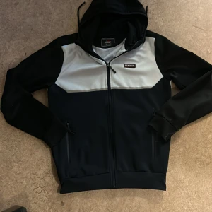 Svart och vit hoddie från McKenzie - Snygg hoddie från McKenzie i svart och vitt med huva och hel dragkedja framtill. Hoddie har två sidofickor med dragkedja och en sportig look. Materialet är polyester och insidan är mjukt fodrad. Perfekt för dig som gillar streetwear.