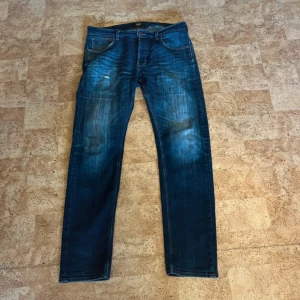 Blå jeans från Lee med raka ben - Säljer ett par klassiska blå jeans från Lee med snyggt slitna detaljer och raka ben. Jeansen har fem fickor, bälteshällor och Lee-logga på bakfickan. Perfekta för dig som gillar en tidlös och avslappnad stil.