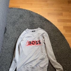 Grå BOSS sweatshirt med röd drakdetalj - Snygg grå sweatshirt från BOSS med svart logga och röda drakdetaljer framtill. Tröjan har rund halsringning och långa ärmar. Perfekt för dig som vill ha en stilren men ändå unik look.