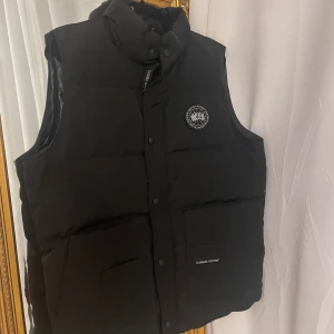 Svart dunväst från Canada Goose - HELT NY & OANVÄND Svart dunväst från Canada Goose i storlek XL med hög krage, tryckknappar och dragkedja. Västen har två stora fickor framtill och klassisk logotyp på bröstet.  lappar sitter kvar med QR som bevis på äkta vara. Pris kan diskuteras över 1200kr