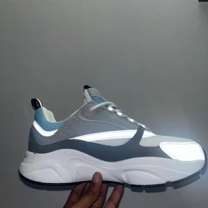 Dior sneakers i grått och blått - Chunky Dior sneakers med vit, grå och ljusblå färg, reflekterande detaljer och meshpaneler. Snygg vågig sula i vitt och svart med blå accent. Skorna har snörning och sportig vibe, perfekt för dig som gillar statement sneakers.