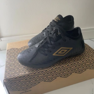 Svarta Umbro fotbollsskor (barn) - Snygga svarta fotbollsskor från Umbro med guldig logga på sidan och tungan. Skorna har snörning och är tillverkade i syntetmaterial med diskret mönster på ovandelen. Perfekta för dig som vill sticka ut på planen med stilrena detaljer.