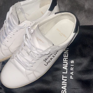 Saint Laurent vita sneakers i skinn - Säljer ett par stilrena vita sneakers från Saint Laurent med svart detalj på hälen och handskriven logga. Jag säljer dessa billigt pga av att mama tog med boxkartongen till återvinningen. Pris kan diskuteras!