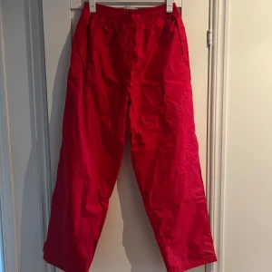 Röda täckbyxor från Airway XXL - Riktigt snygga röda täckbyxor från Airway i storlek XXL. Byxorna har elastisk midja, raka ben och praktiska fickor. Perfekta för kalla dagar när du vill hålla dig varm och ändå se cool ut. Materialet är tåligt och vattenavvisande syntet.