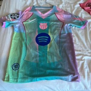 Barça x Nike Lamine Yamal fotbollströja - Säljer en limited edition FC Barcelona x Nike fotbollströja med Lamine Yamal och nummer 19 på ryggen. Tröjan har coola pastellfärger i turkos, rosa, ljusblå och gul, med mönstrade detaljer och tryck från Spotify. Kortärmad och tillverkad i lätt, ventilerande polyester.